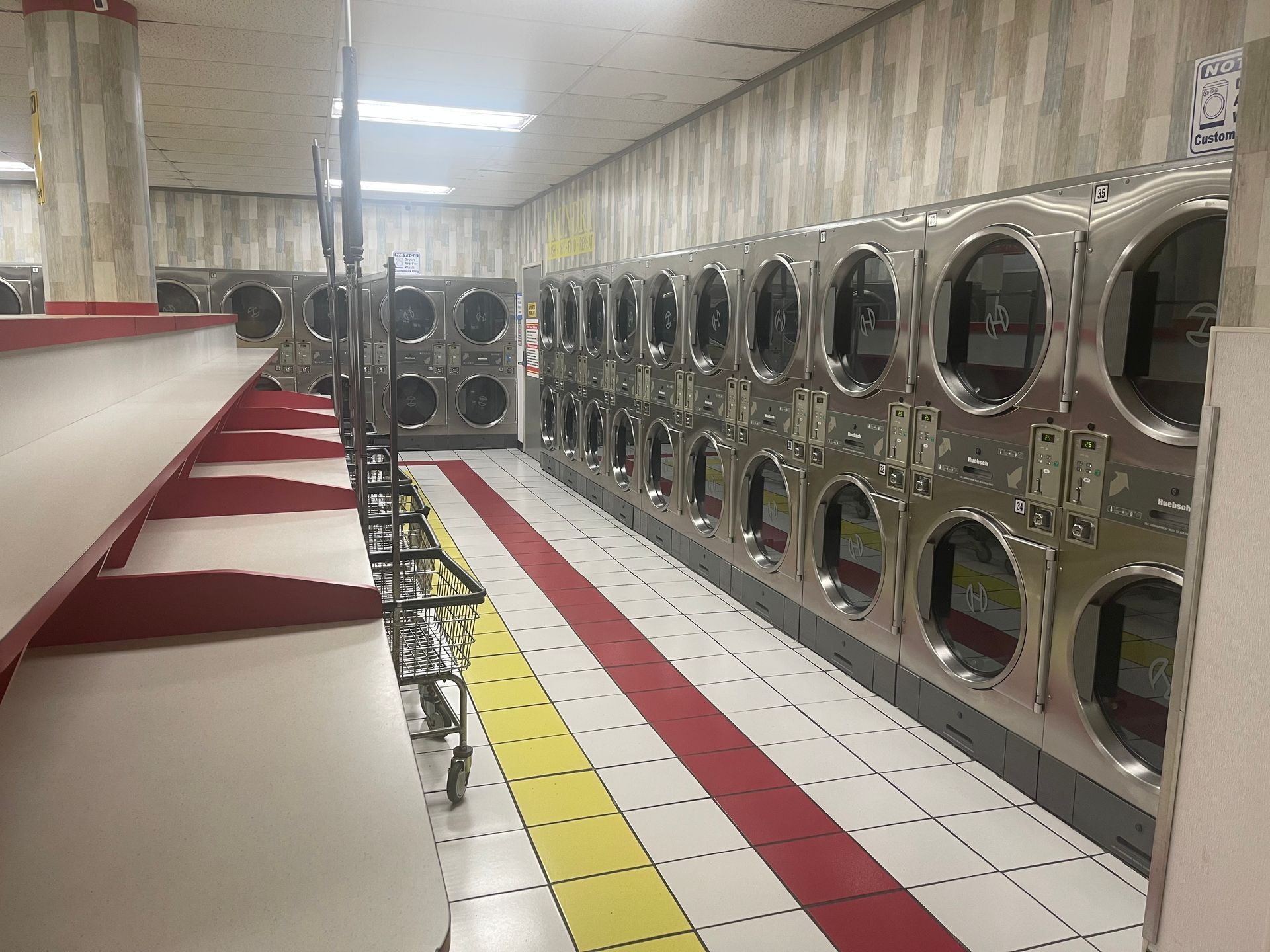 A laundromat with rows of silver dryers and folding tables, red and yellow floor accents.