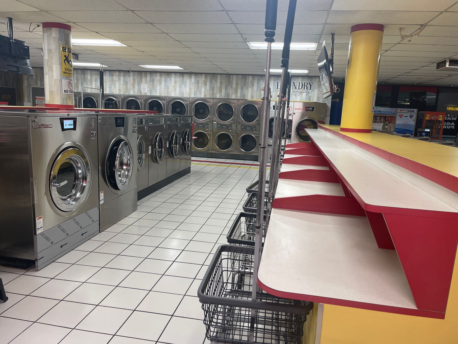Laundromat interior with rows of washers and dryers. A red and white folding table with baskets.
