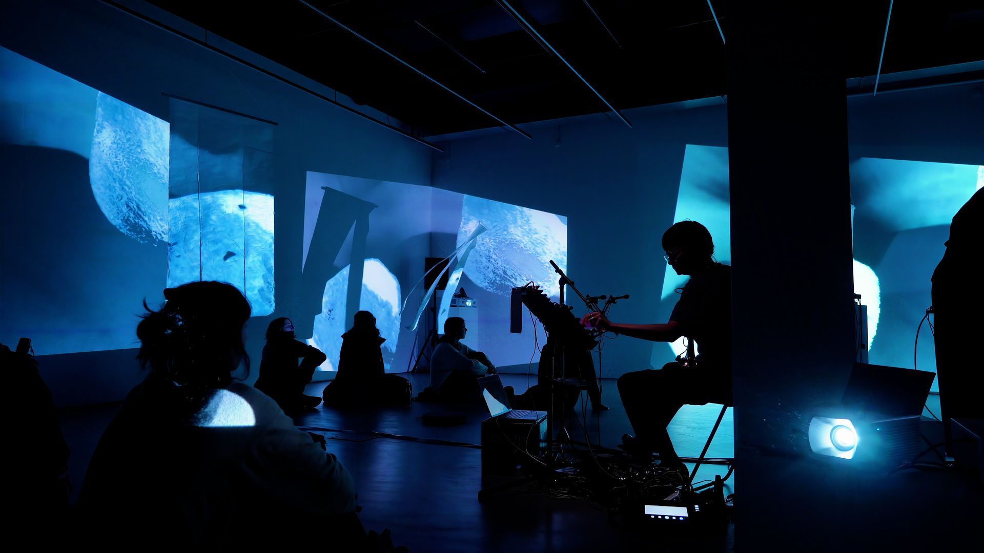 A silhouetted audience watches a live musical performance in a darkened room with blue, abstract projections on the wall.