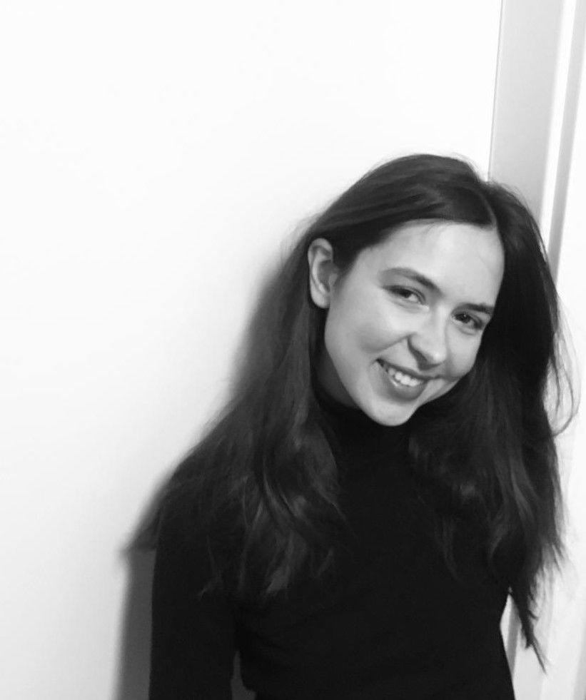 A smiling person with long dark hair wears a black turtleneck while standing against a white wall.