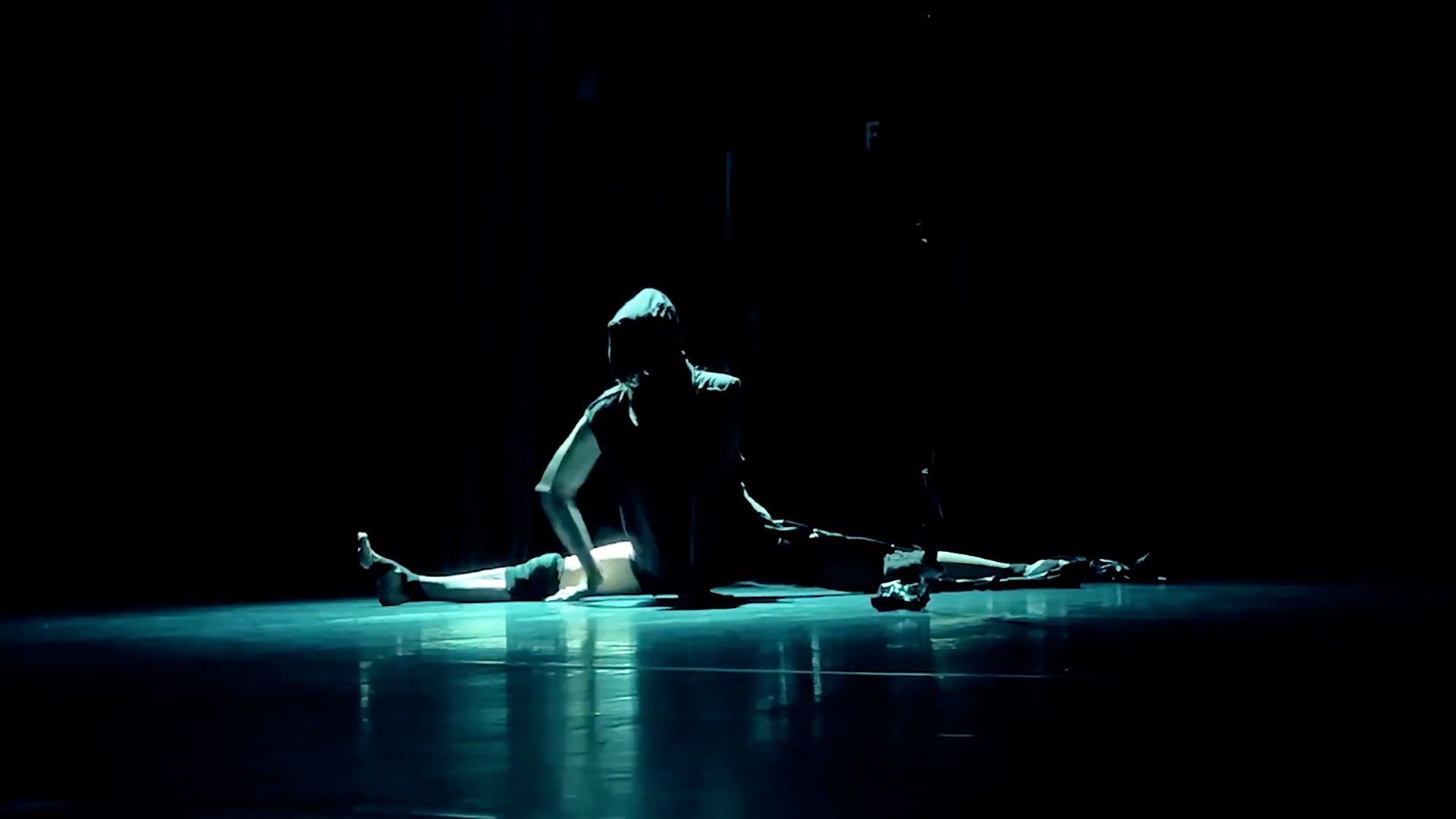 A dancer performs a center split on a dark, dimly lit stage, illuminated by a pool of cool blue light.