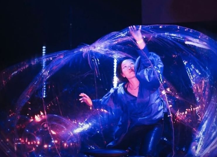A performer in a blue jacket sits inside a large, transparent bubble, reaching upward amidst glowing purple lights.