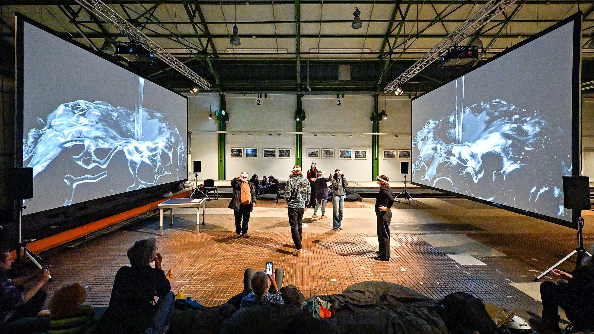 People in a large hall watch two giant screens displaying flowing water, with a stage area between them.