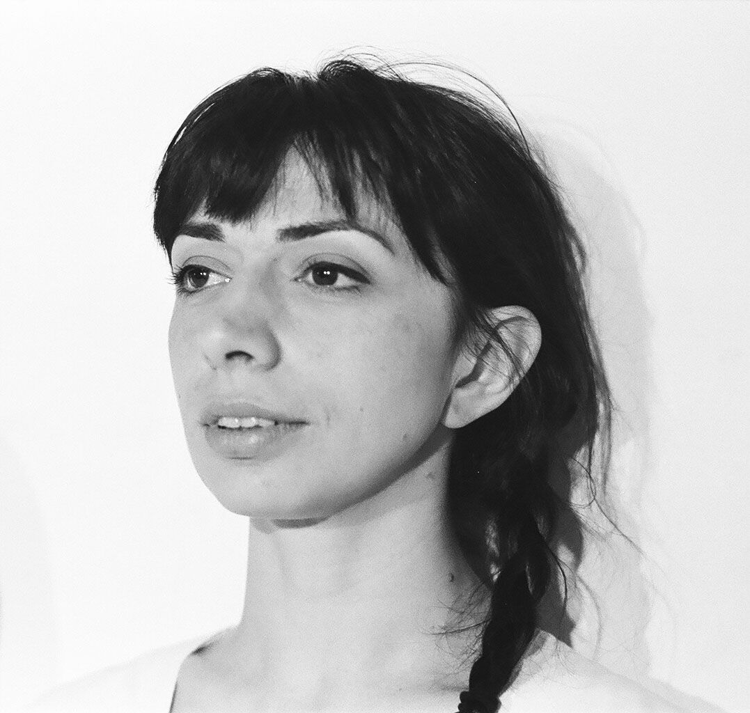 A black-and-white portrait of a person with bangs and a side braid, looking slightly to the side against a white wall.