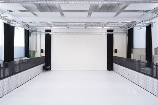 Empty performance hall with white walls and floors, black curtains, and a gridded metal ceiling structure.