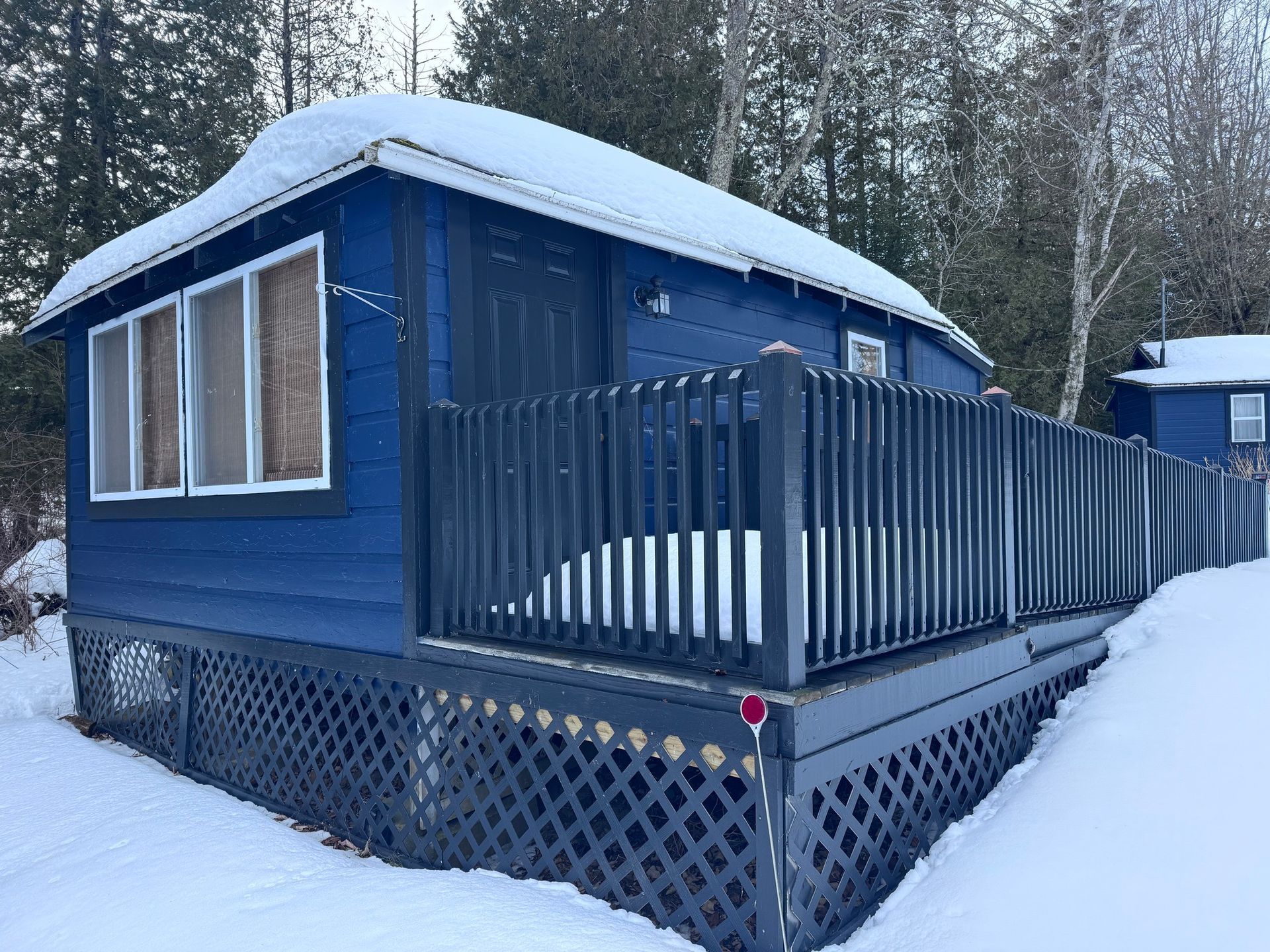 Blue cabin with snow-covered roof and porch railing in a snowy wooded setting