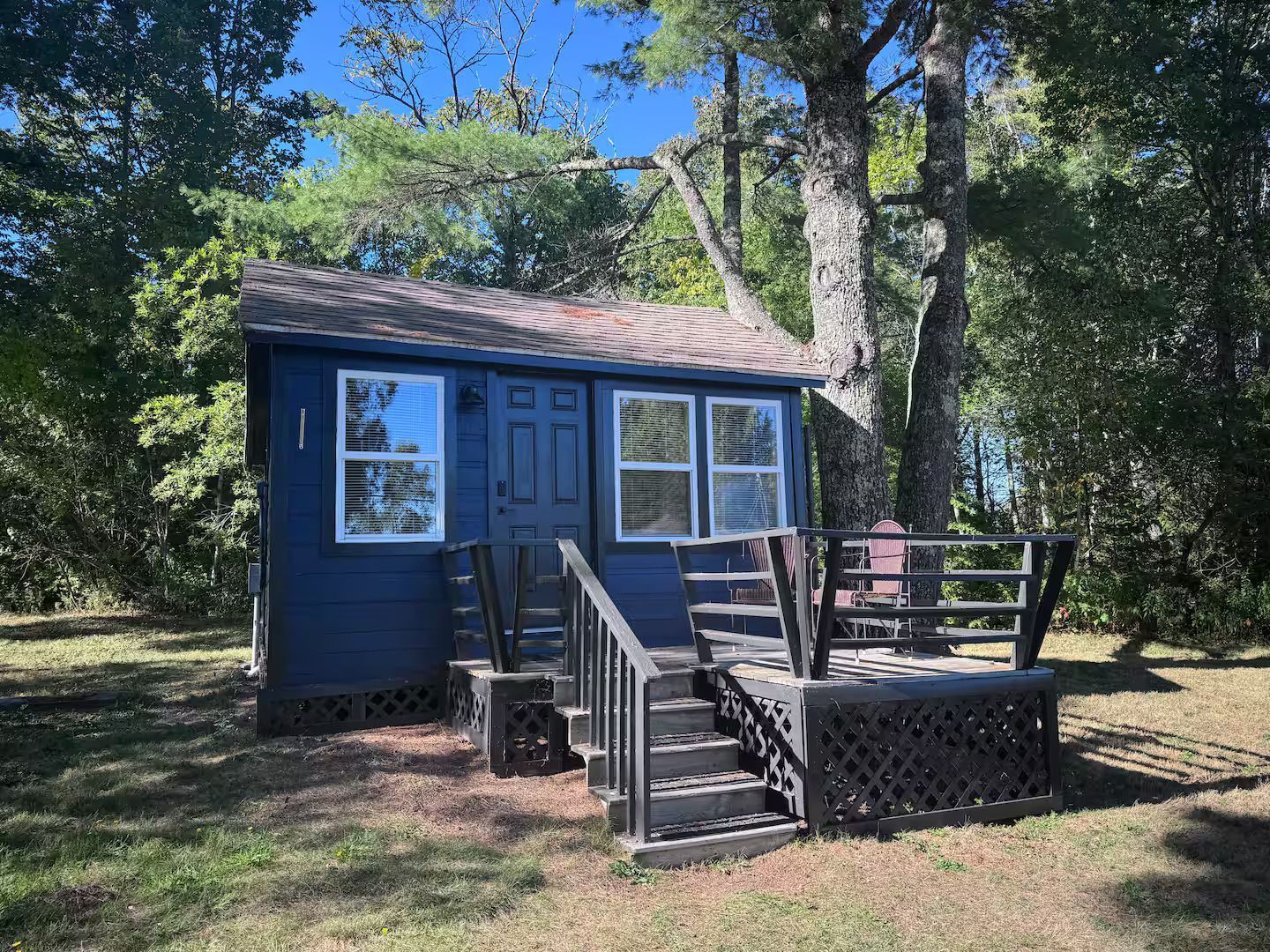 A small, dark blue cabin with a brown roof and a wooden deck, set among lush green trees on a sunny day.