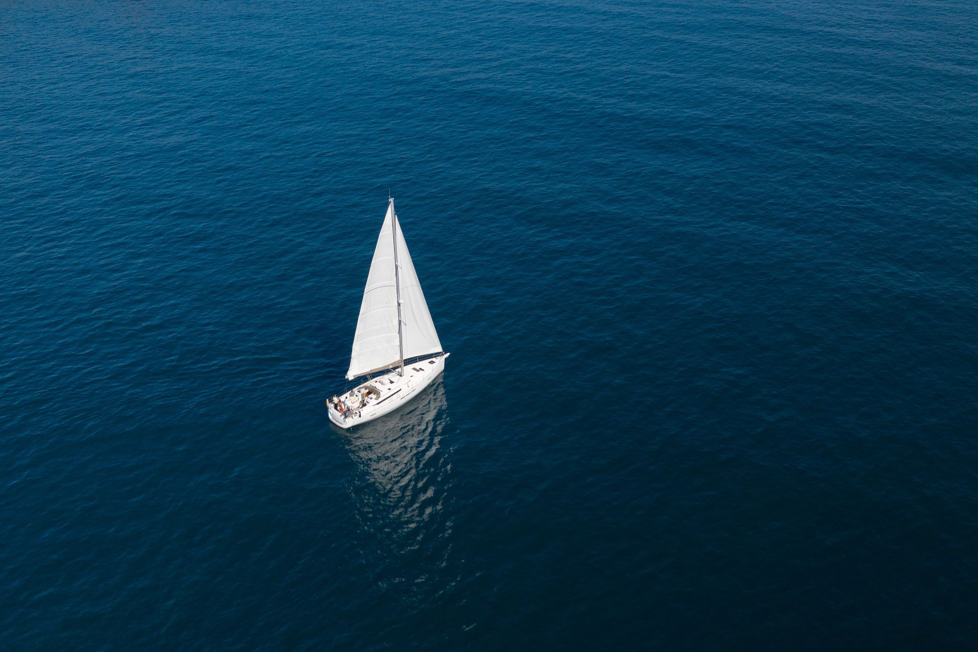 A sailboat with a white sail travels across deep blue, calm ocean water, viewed from directly above.