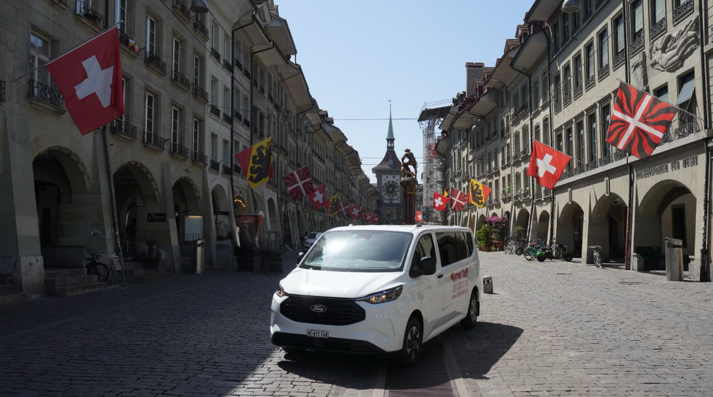 Ein weißer Lieferwagen auf einer mit Schweizer Flaggen gesäumten Kopfsteinpflasterstraße im historischen Stadtzentrum