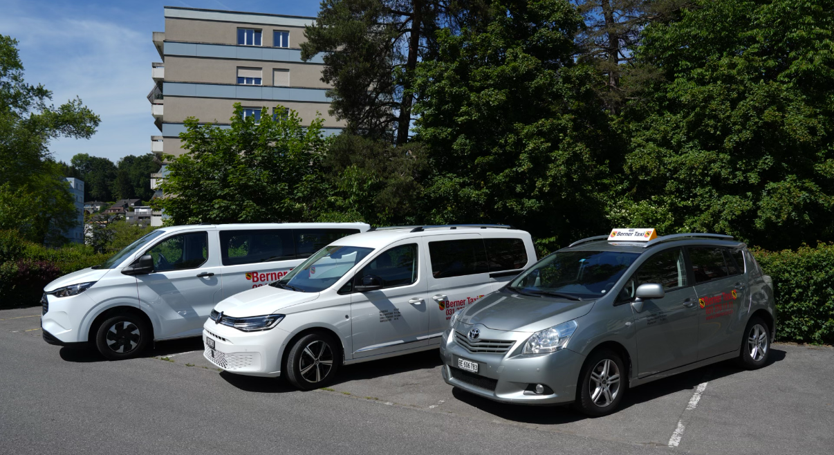 Drei geparkte Autos auf einem Parkplatz, im Hintergrund ein Gebäude und Bäume.