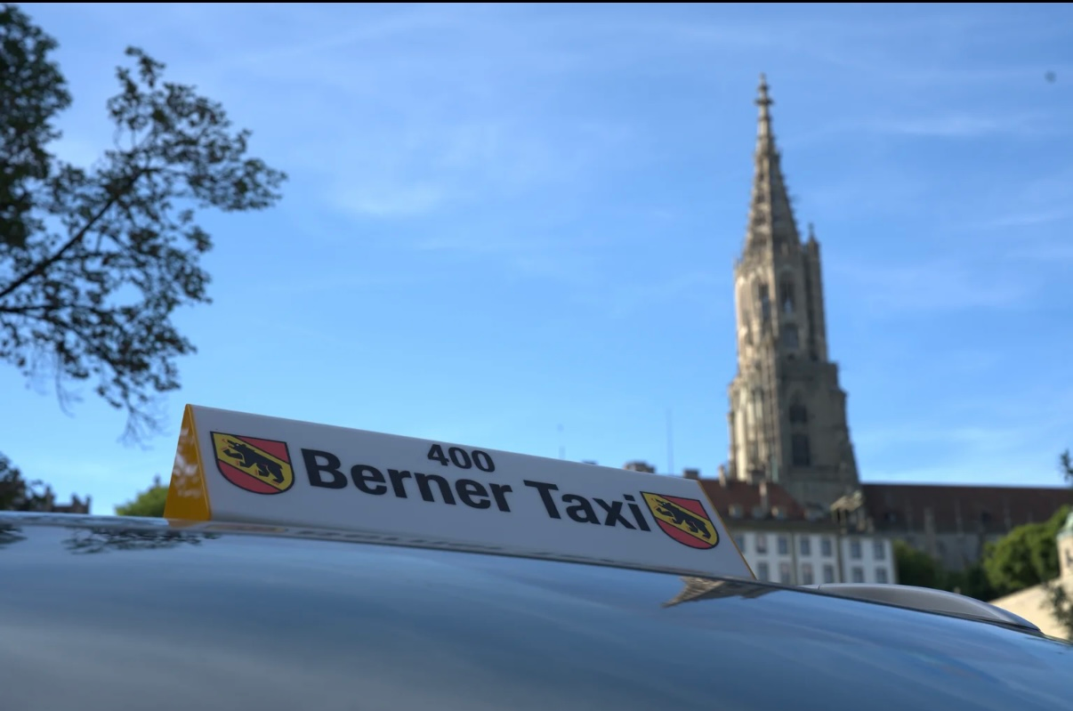 Das Dachschild von Berner Taxi vor einem hohen Kirchturm unter blauem Himmel
