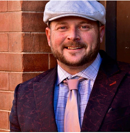 A man wearing a hat and tie is smiling in front of a brick wall