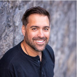 A man with a beard is smiling for the camera while leaning against a wall.