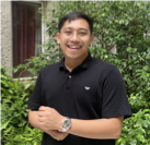 A man wearing a black shirt and a watch is standing in front of a bush.