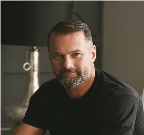 A man with a beard is wearing a black shirt and looking at the camera.