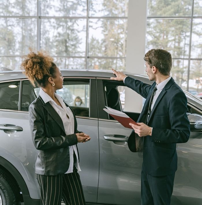 Two businesspeople discussing a silver car in a bright showroom, one holding a clipboard.
