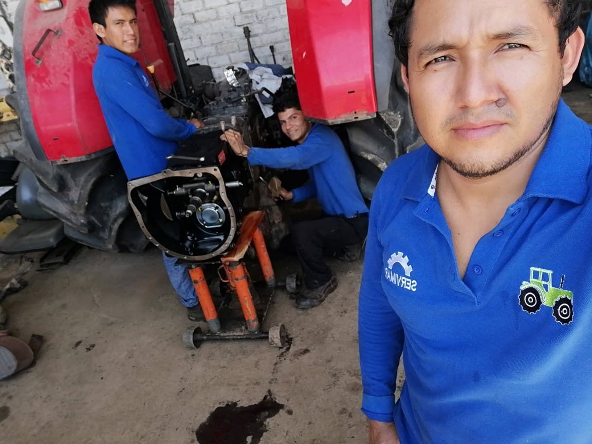 Tres mecánicos trabajando en un tractor, uno de ellos tomándose una selfi. Llevan camisas azules en un taller.