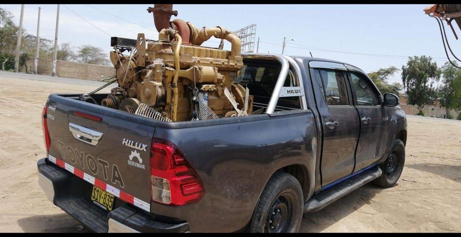 Caja de camioneta Toyota Hilux color gris oscuro cargada con un gran motor Caterpillar color beige.