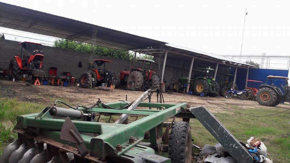 Tractores y maquinaria agrícola aparcados bajo un refugio en una granja, con una grada de discos verde en primer plano.