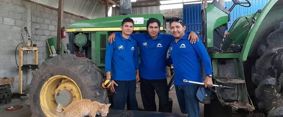 Tres hombres con camisas azules posan con un tractor John Deere en un garaje. Un gato está sentado cerca de la llanta.