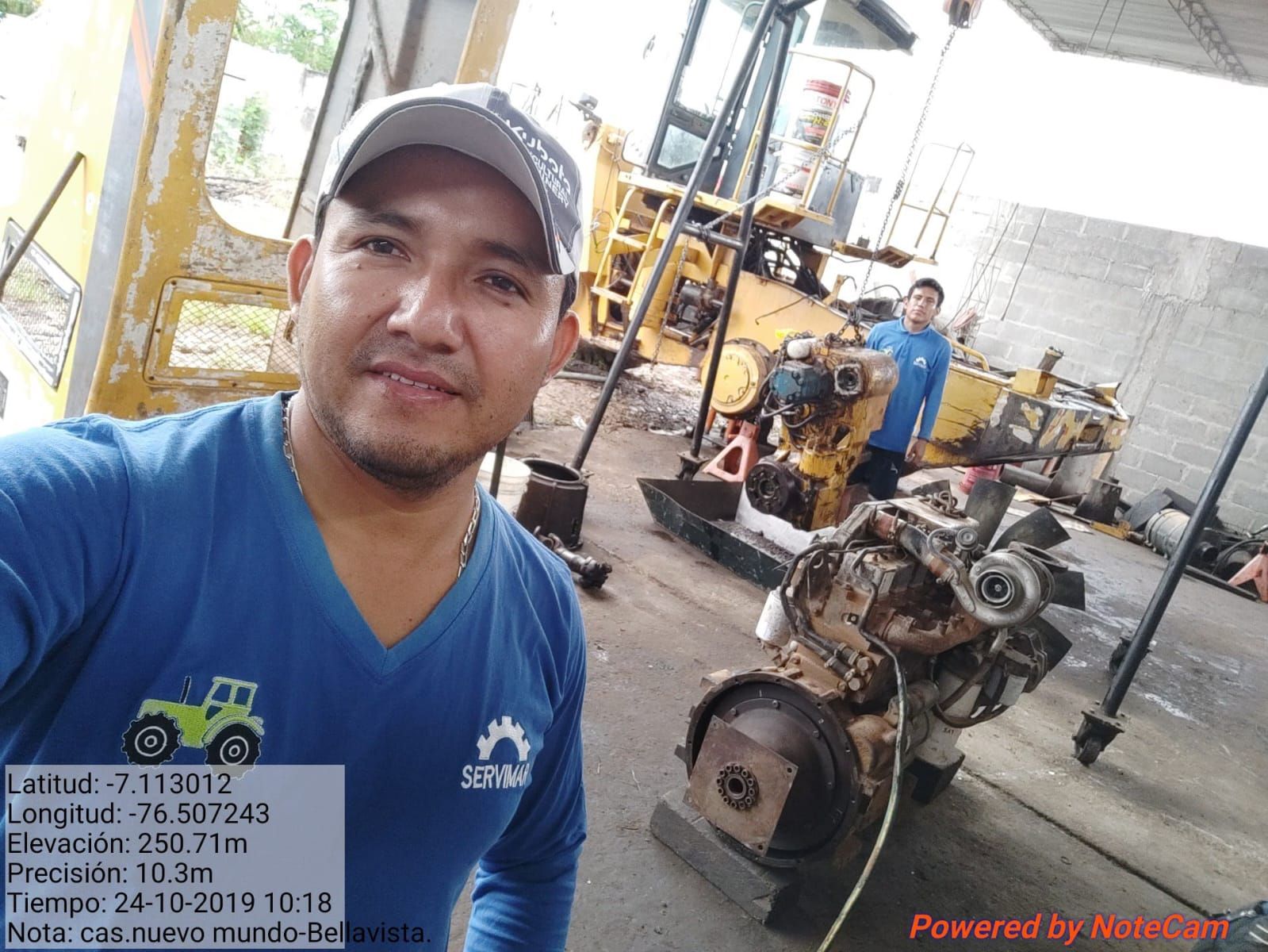 Un hombre con camisa azul y gorra se toma un selfi en un taller con maquinaria pesada. Hay otra persona al fondo.