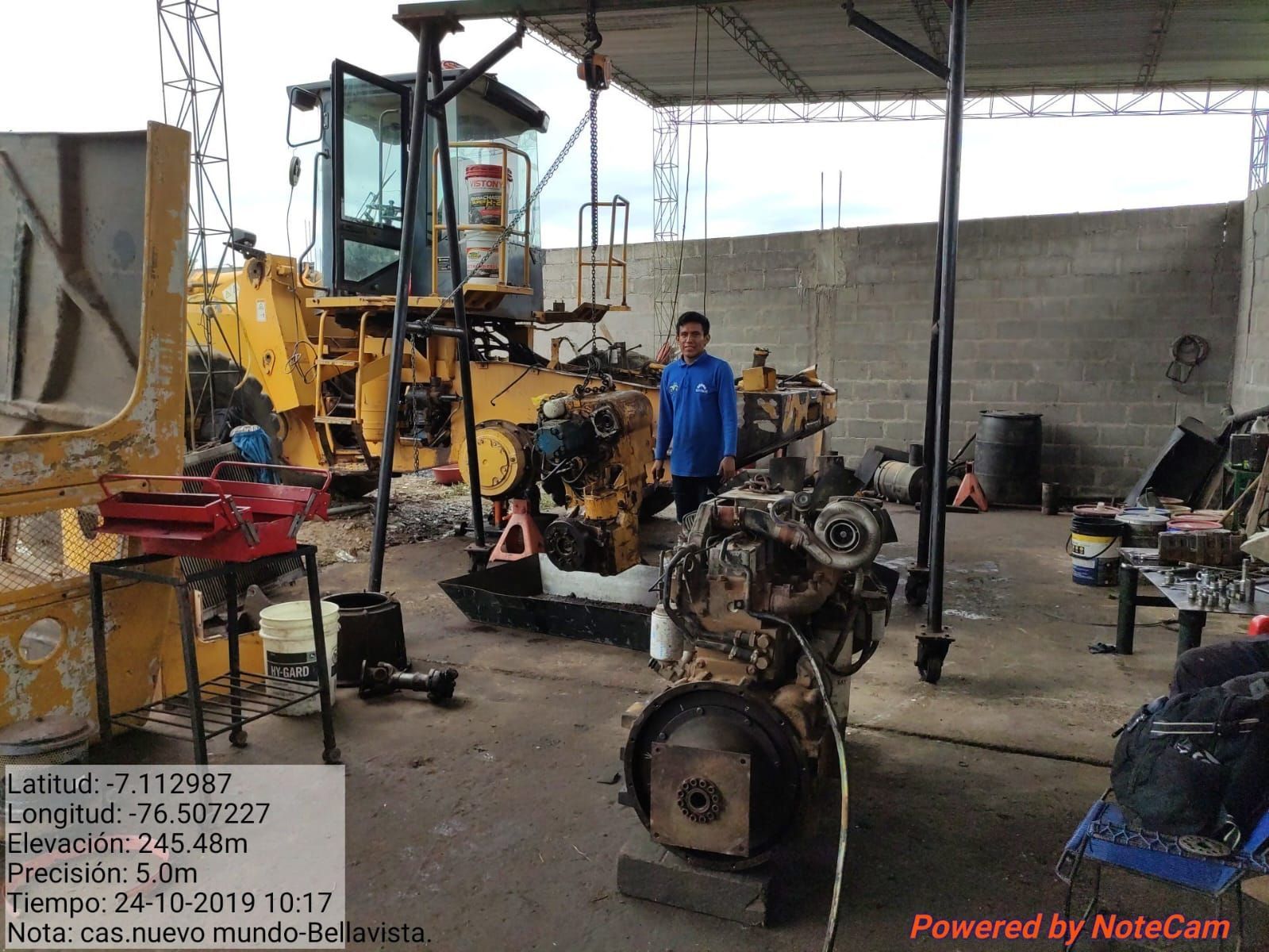 Un hombre se encuentra en un taller de reparación con maquinaria pesada y motores. Se muestran las coordenadas y la fecha.