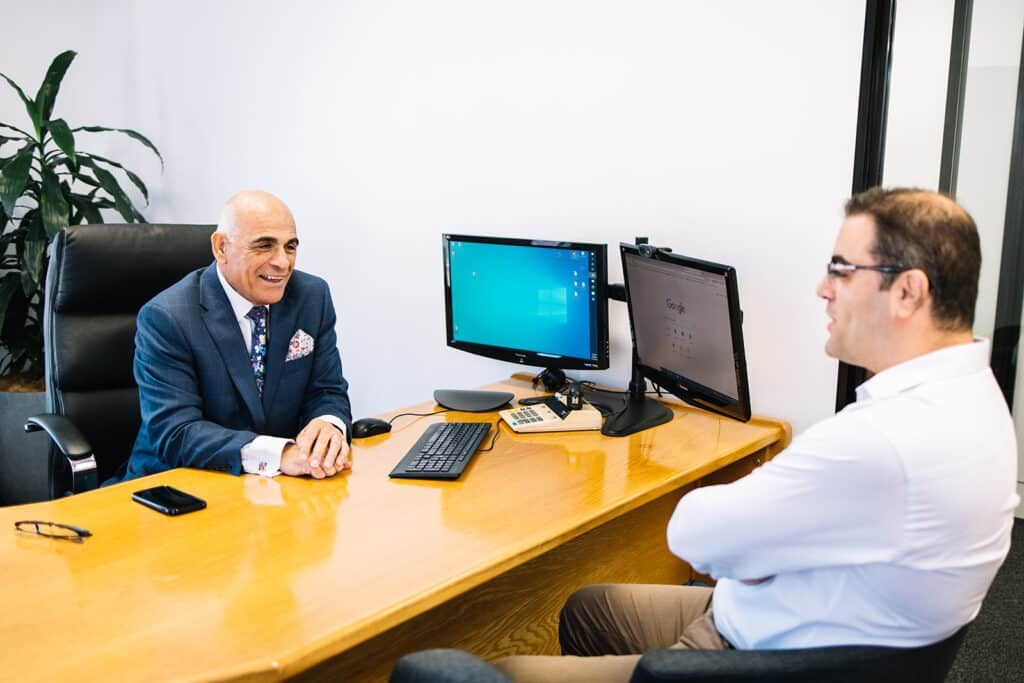 Two Men Are Sitting At A Desk Talking To Each Other — The Garis Group  In Hamilton, NSW 