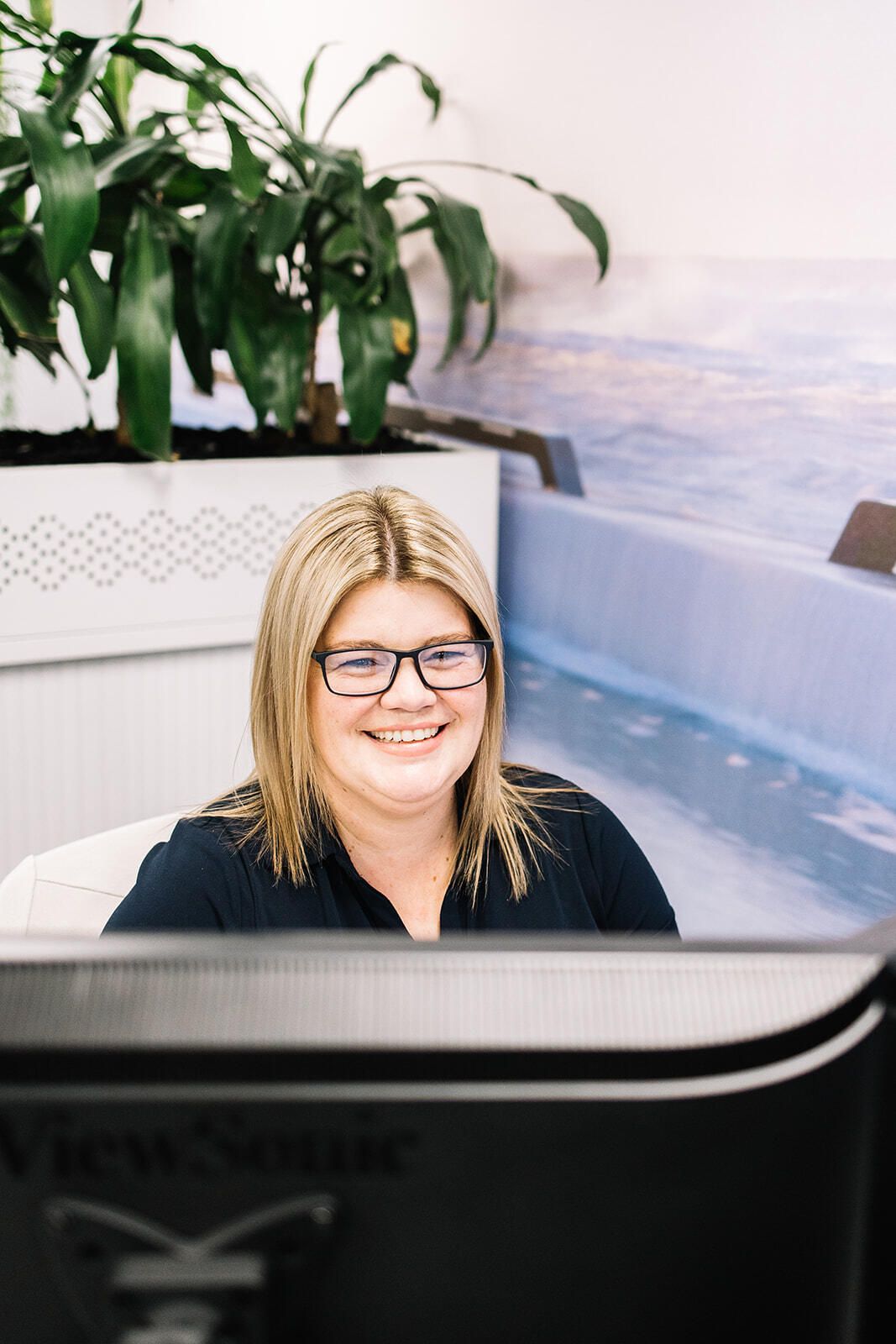 A Woman Wearing Glasses Is Smiling While Sitting In Front Of A Computer Monitor — The Garis Group In Sydney, NSW