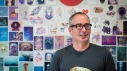 A Man Wearing Glasses Is Standing In Front Of A Wall Covered In Pictures— The Garis Group  In Hamilton, NSW 
