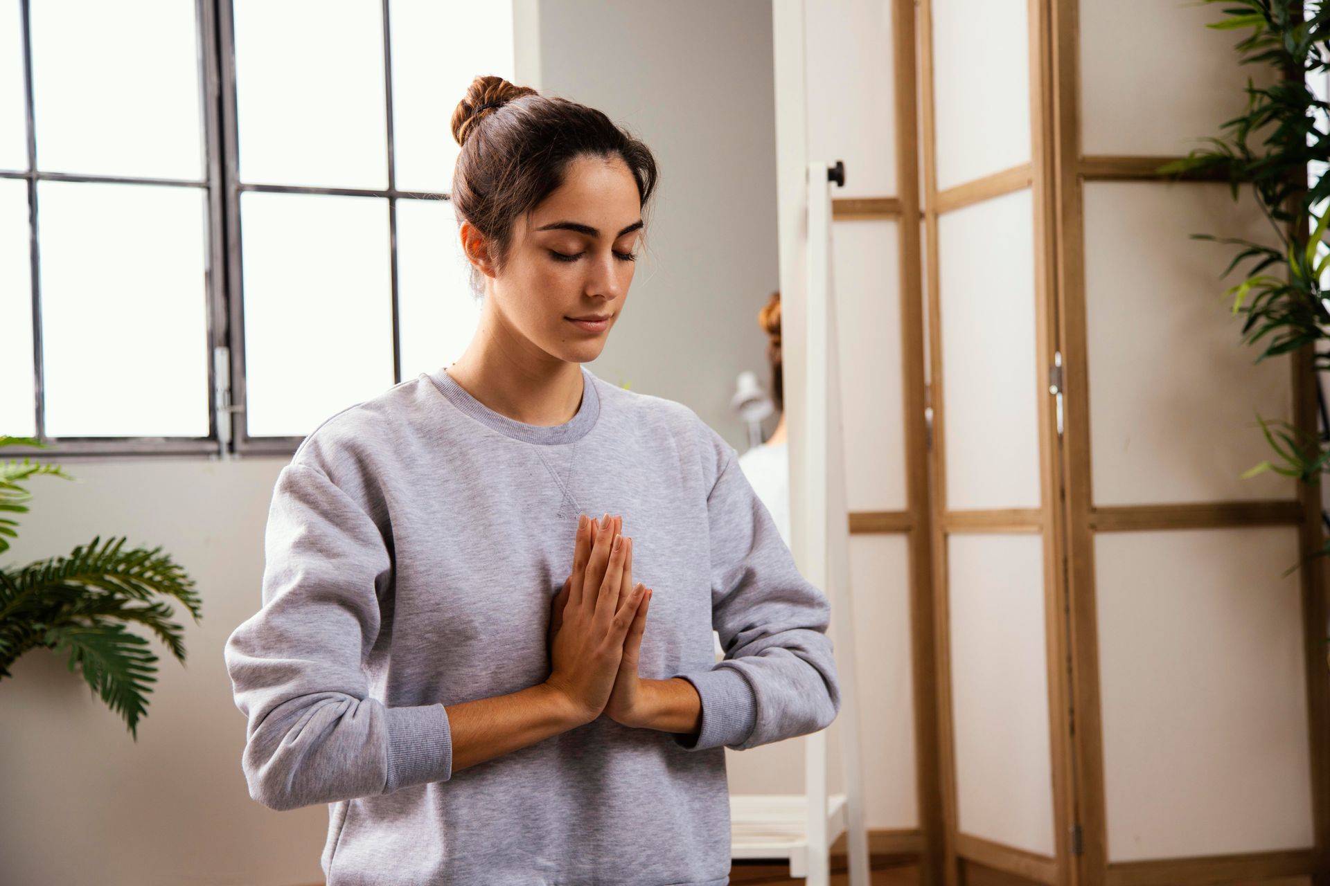 Woman in gray sweater meditating indoors with hands in prayer position.