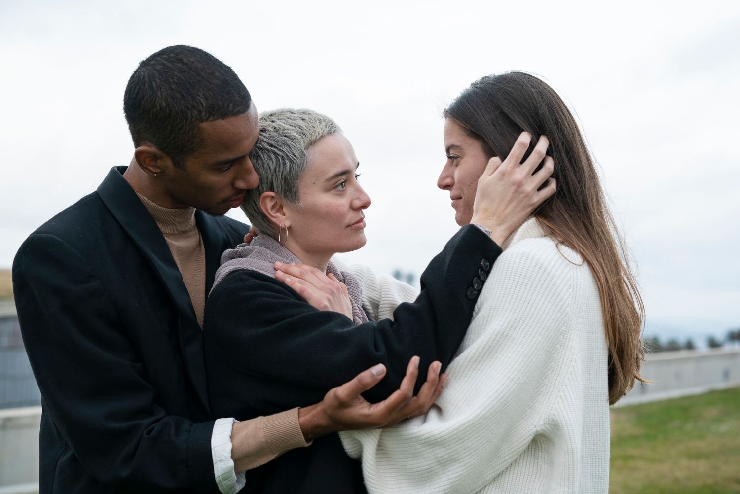 Three people standing outdoors, one gently comforting another by touching her face.