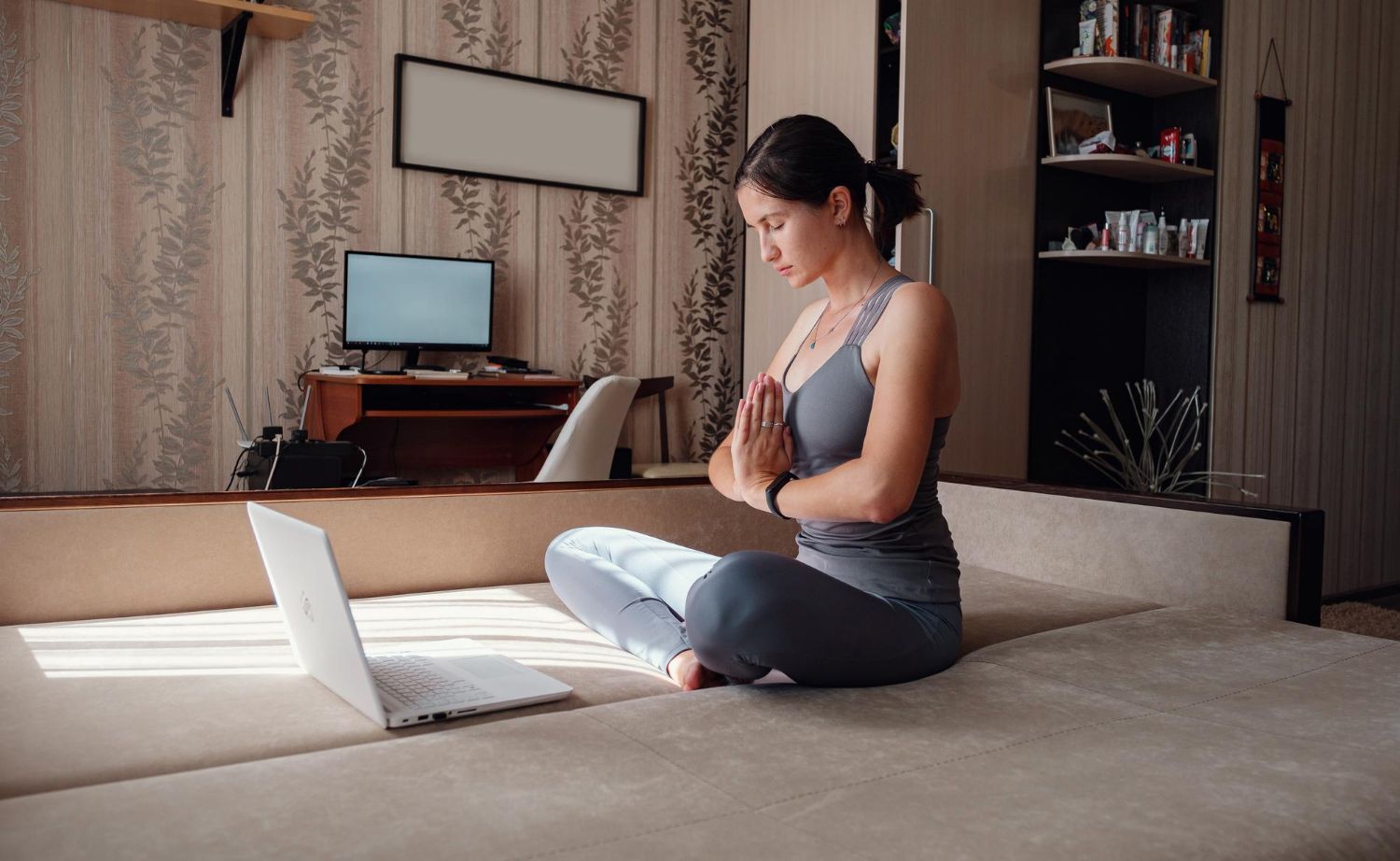 Woman in workout clothes meditates on a sofa, hands in prayer position, laptop open.