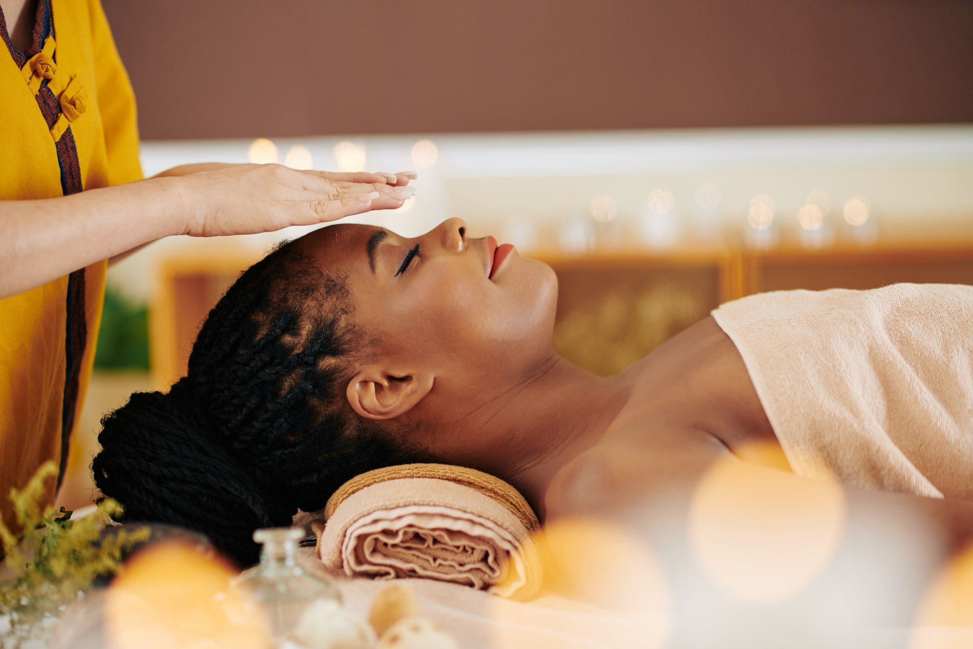 Woman receiving energy healing; practitioner's hand above her head. She is relaxed, eyes closed, in a spa-like setting.