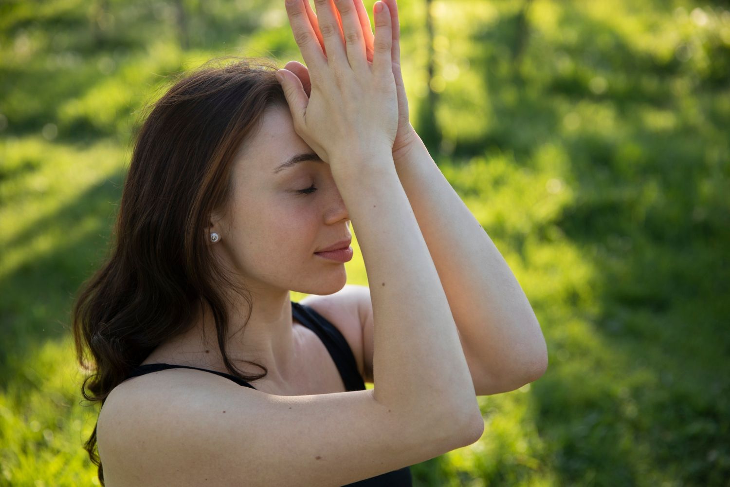 Person in a black top with arms raised, eyes closed, standing outdoors in green sunlight