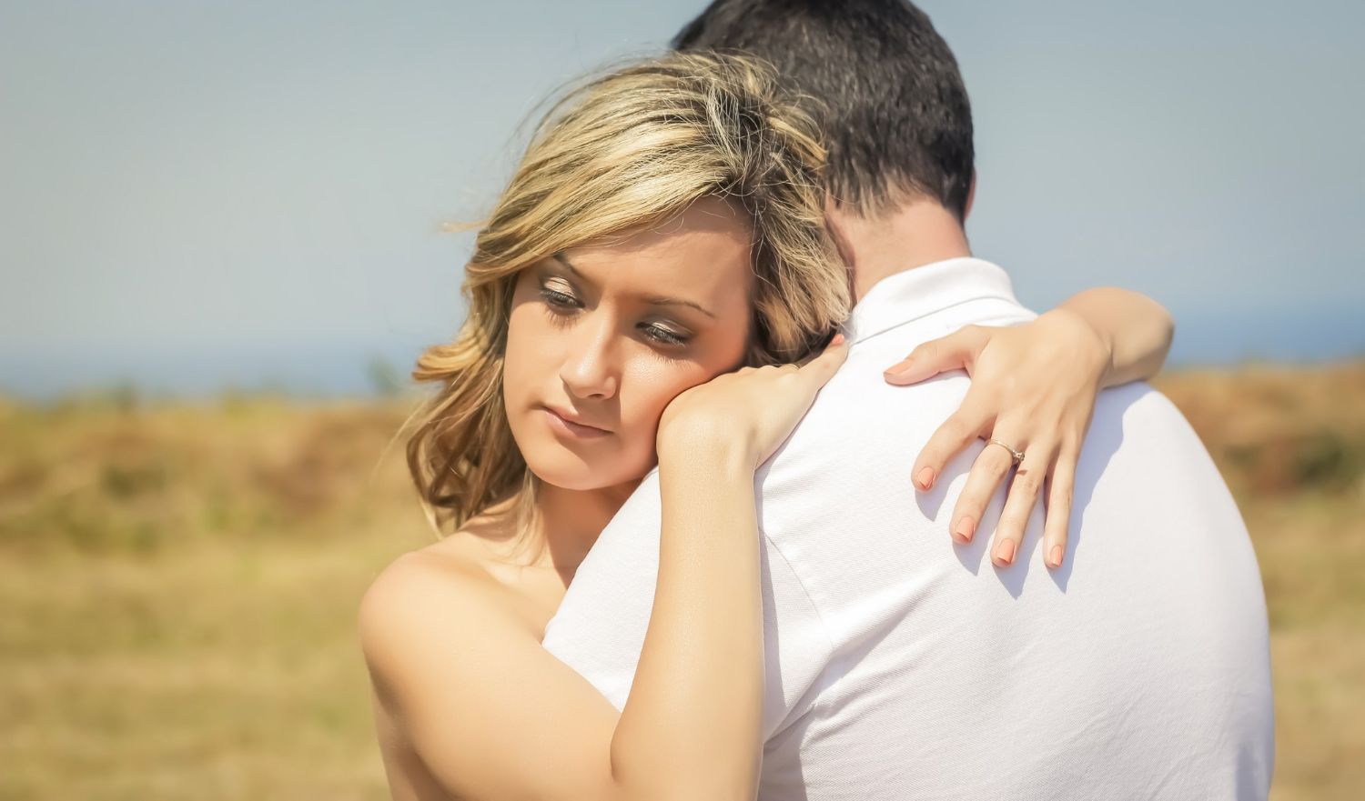 A woman in a white dress hugs a man outdoors in a field, resting her head on his shoulder.