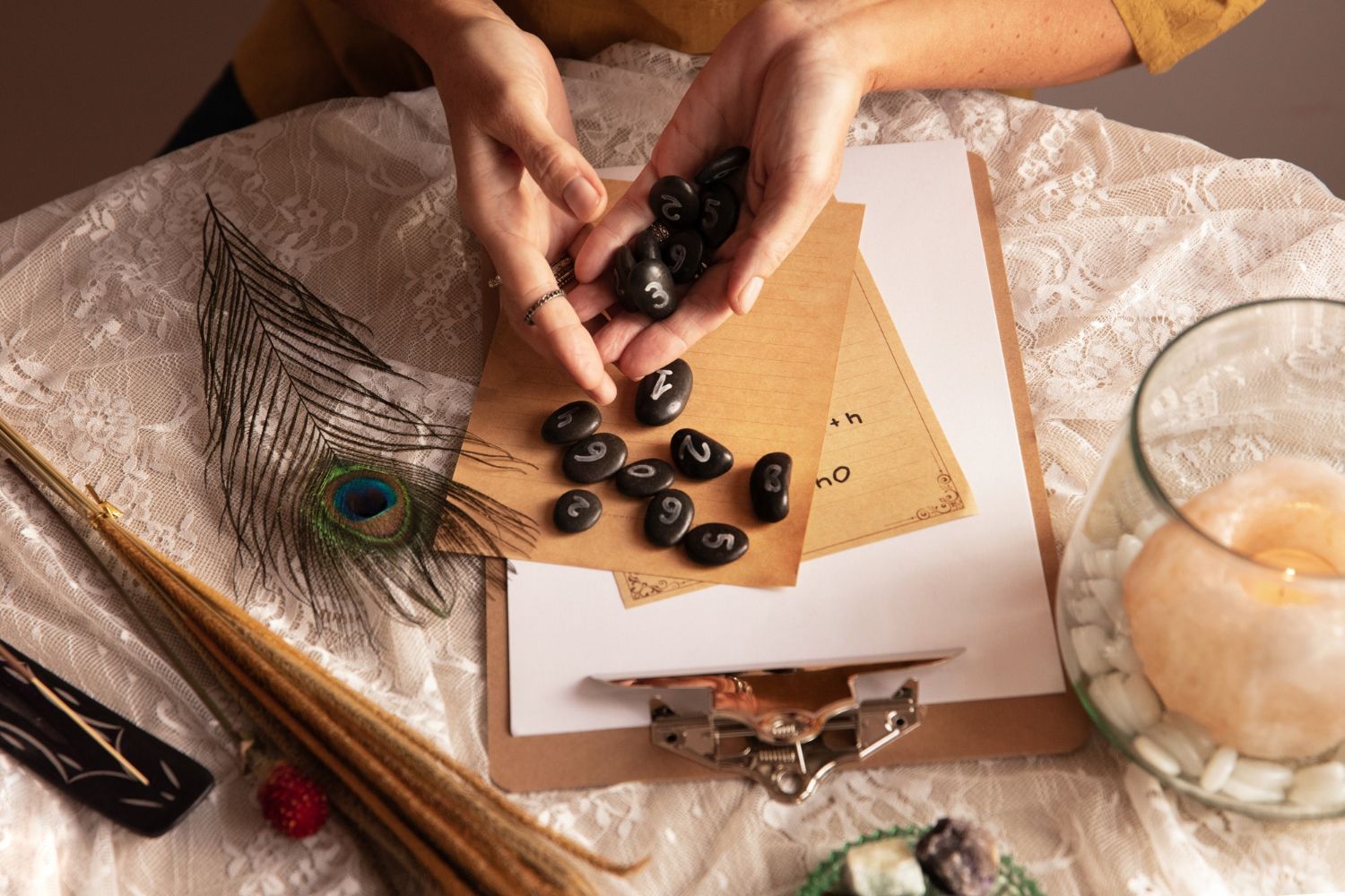 Hands arranging black stones on paper beside a feather, notebook, and candle on a soft fabric surface
