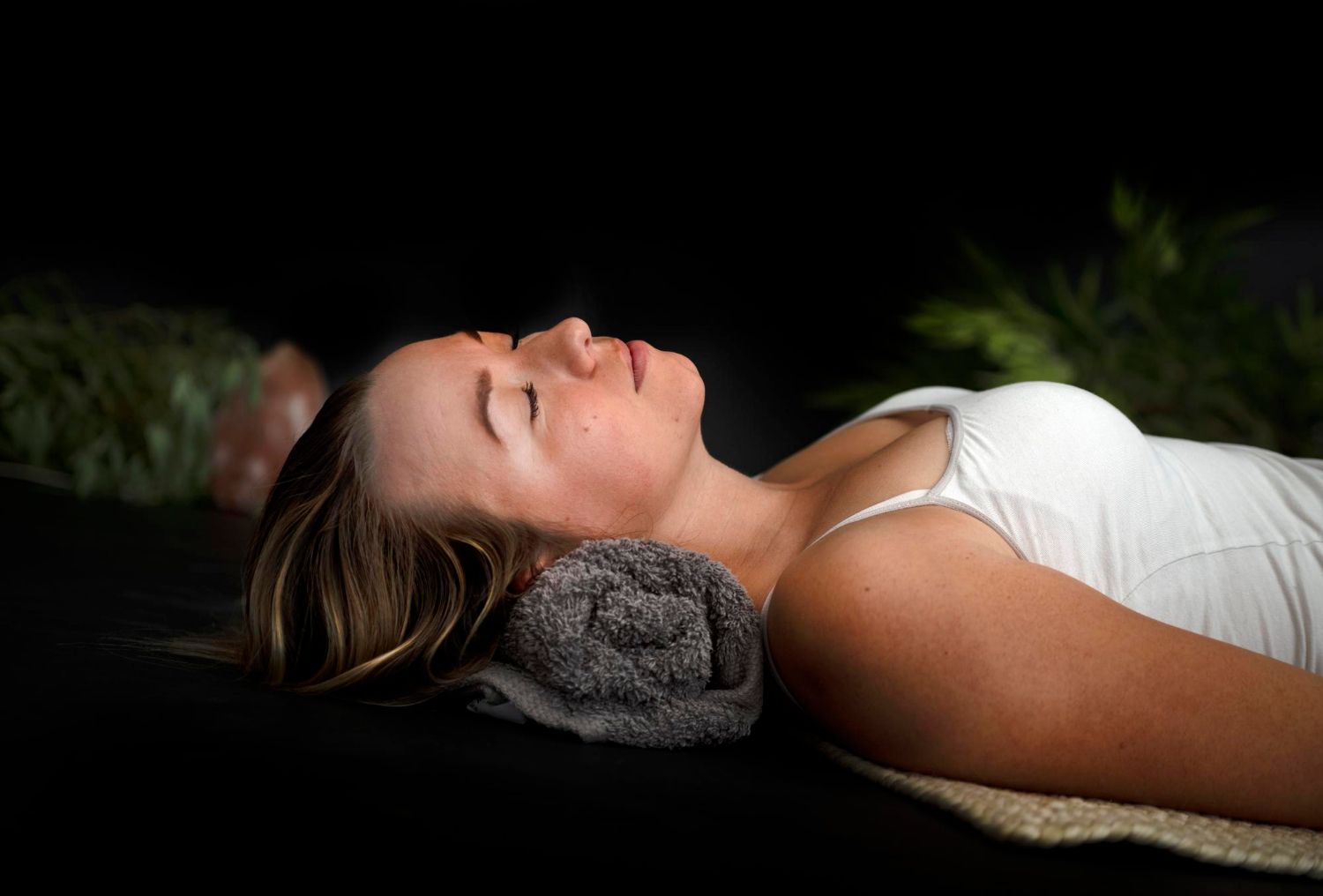 Woman laying down, eyes closed, resting on a rolled towel, in a dark setting, with greenery.