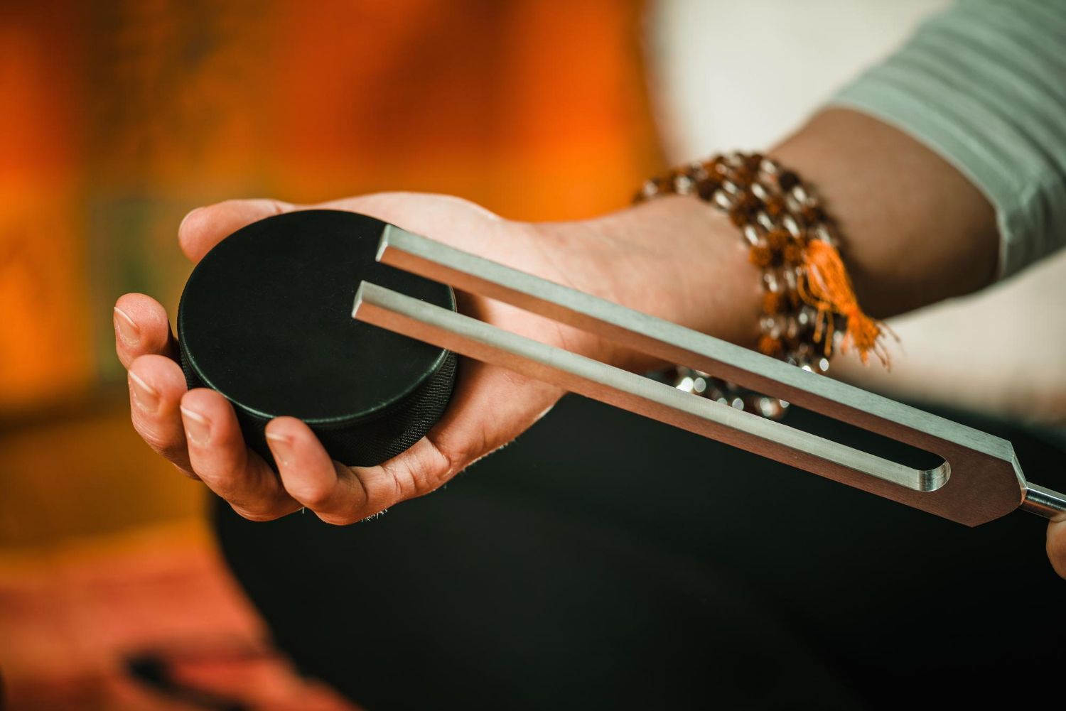Person holding a black tuning fork over a hand, with bracelets on the wrist and a warm orange background