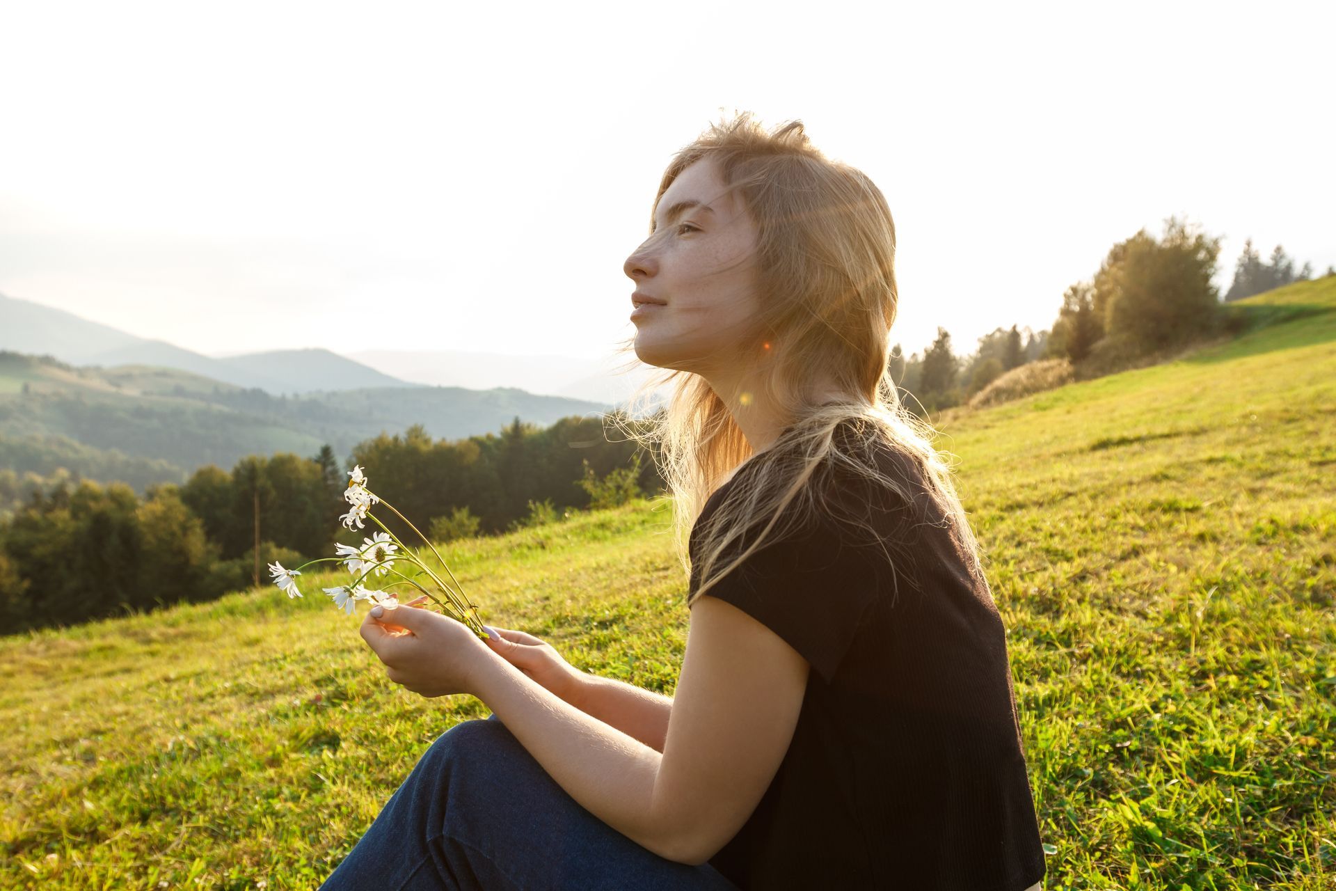 Woman sits on a grassy hillside, eyes closed, holding white flowers, with mountains in the background, illuminated by sunlight.