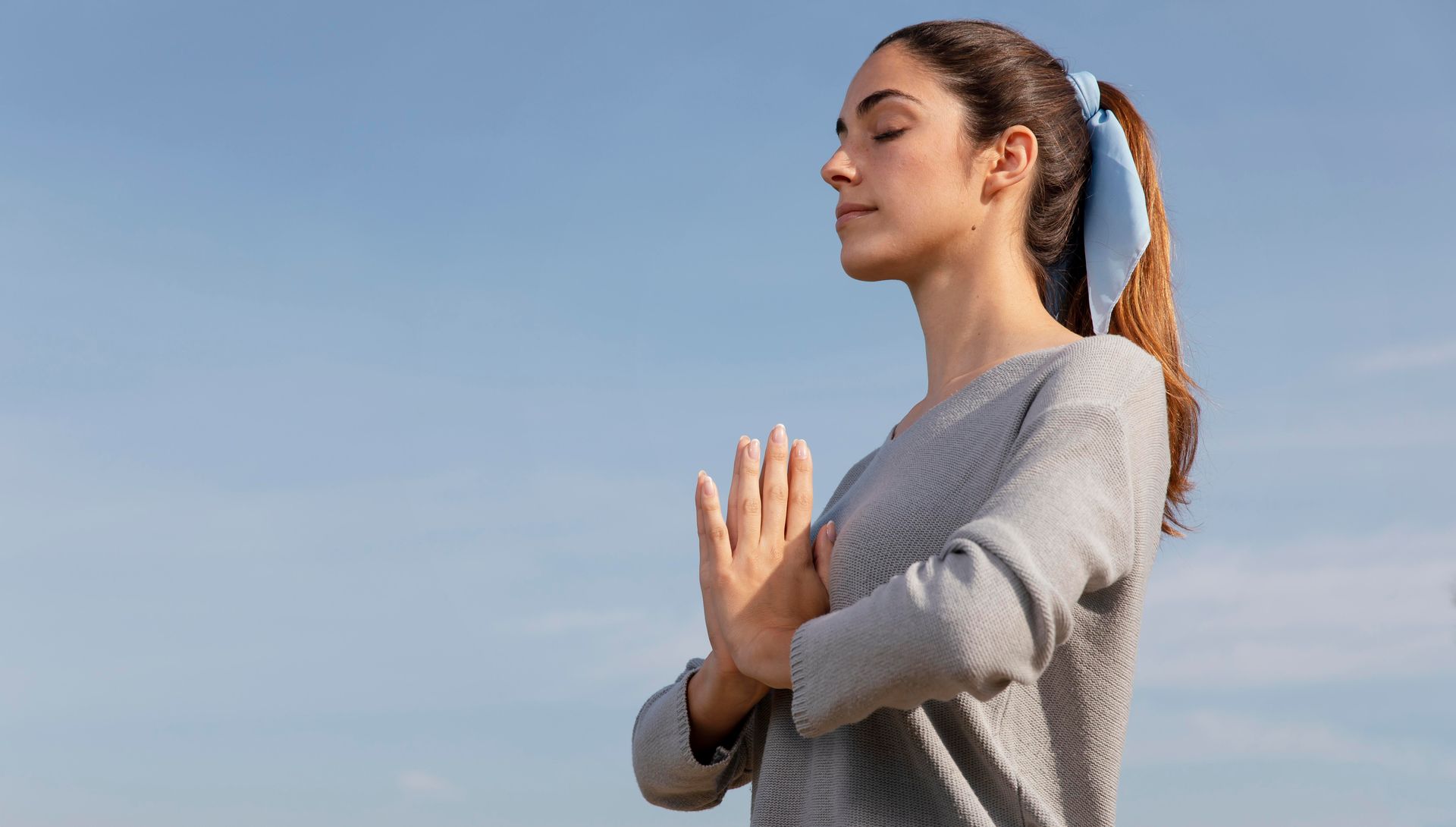 Woman outdoors with eyes closed, hands in prayer position, against a blue sky. She wears a gray sweater and has her hair tied back.