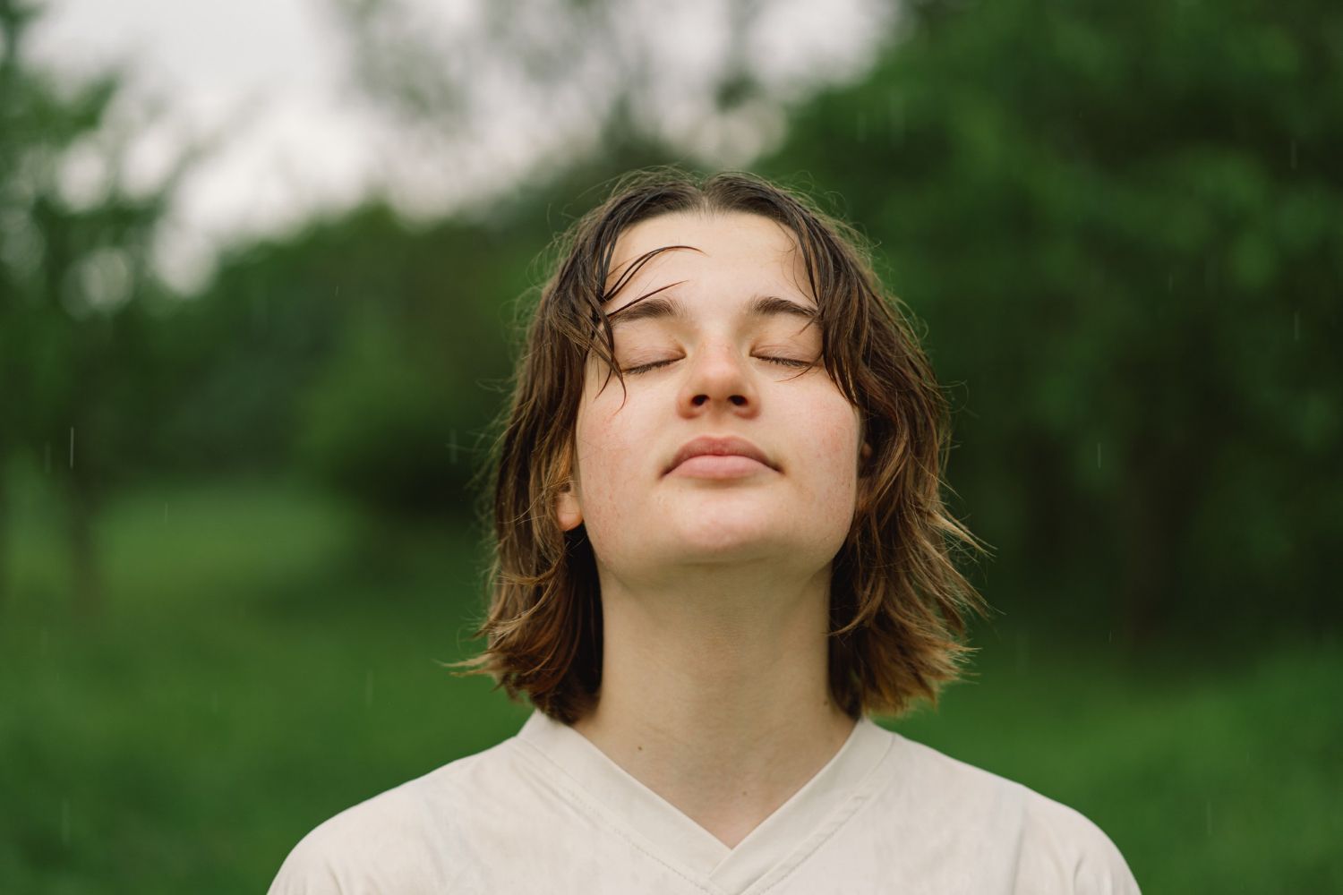 Person with closed eyes standing outdoors in a green park, wearing a light shirt.