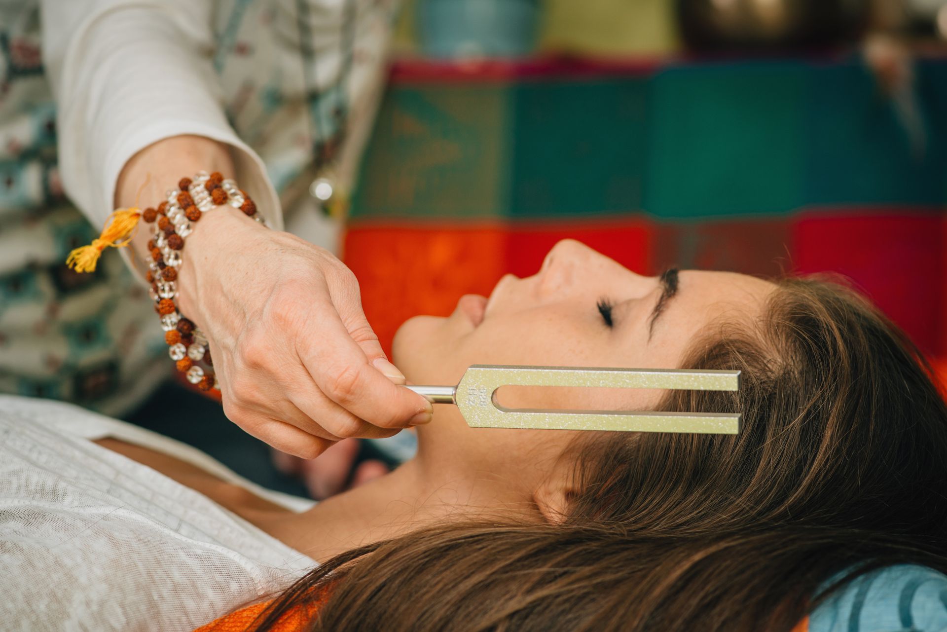 Woman receiving sound healing with a tuning fork held near her face. Someone's hand holding the fork; soft lighting.