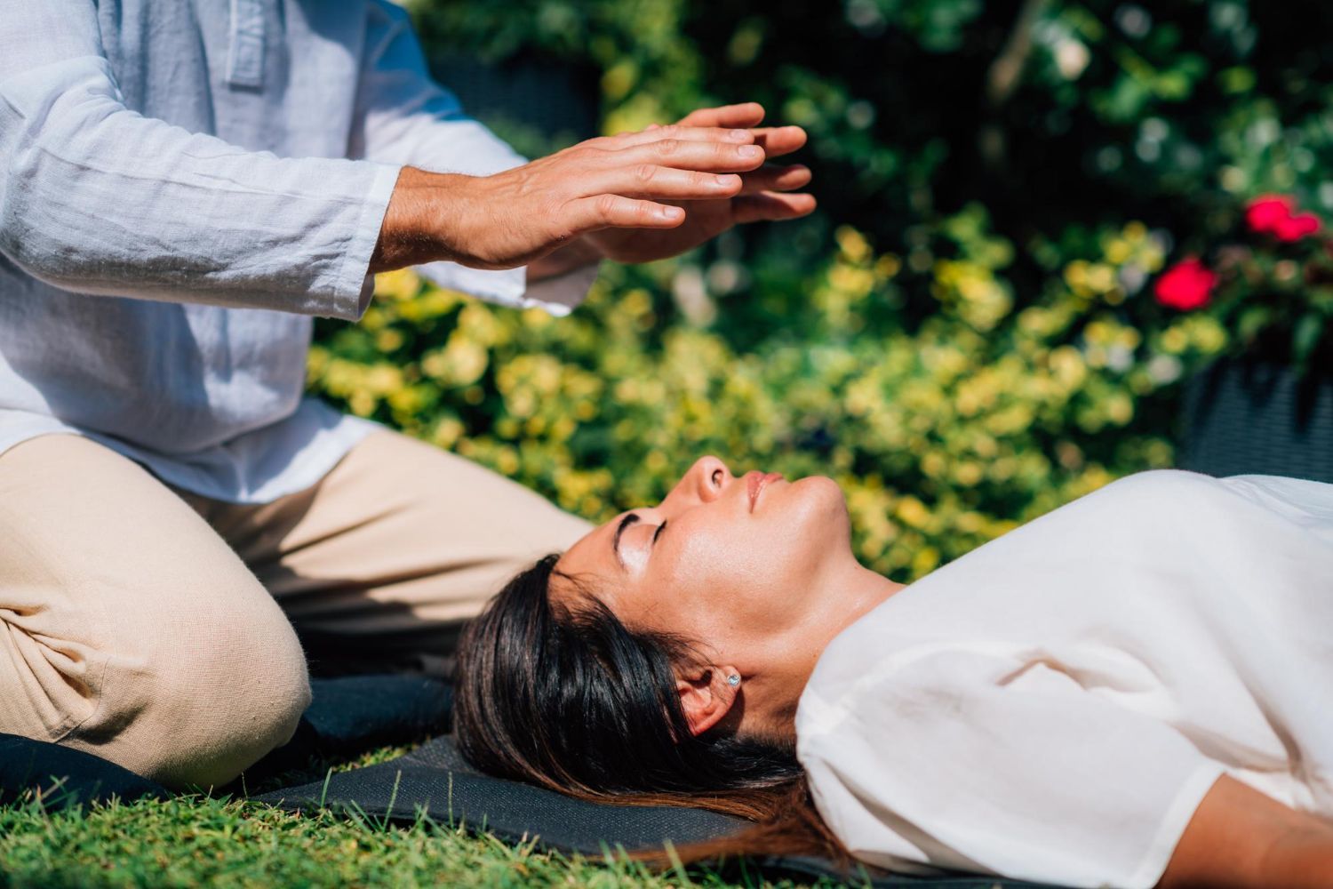Person lying on grass while another kneels nearby, hands raised in a healing gesture in a garden