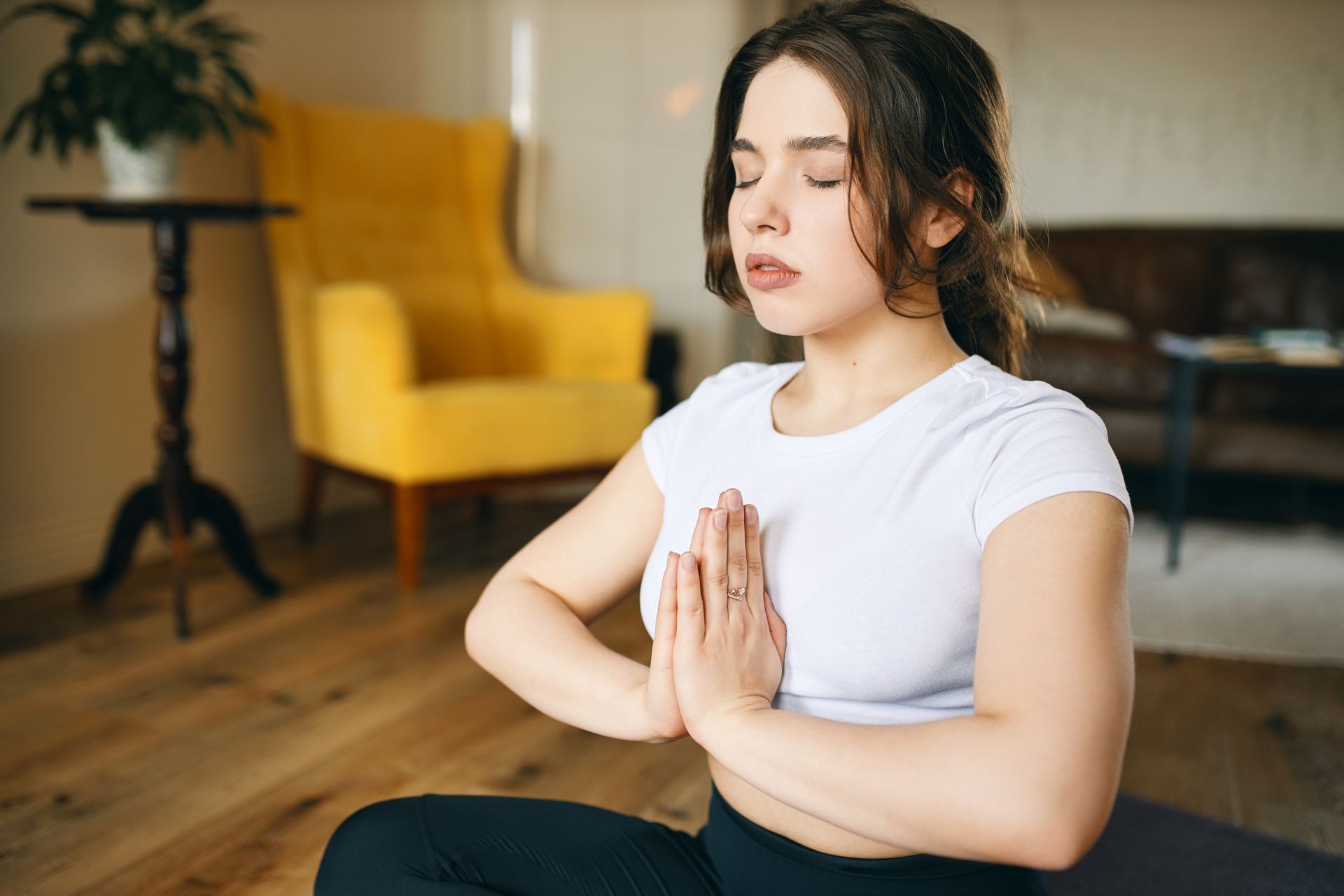 Woman in a white shirt and black leggings meditating with hands together, eyes closed.