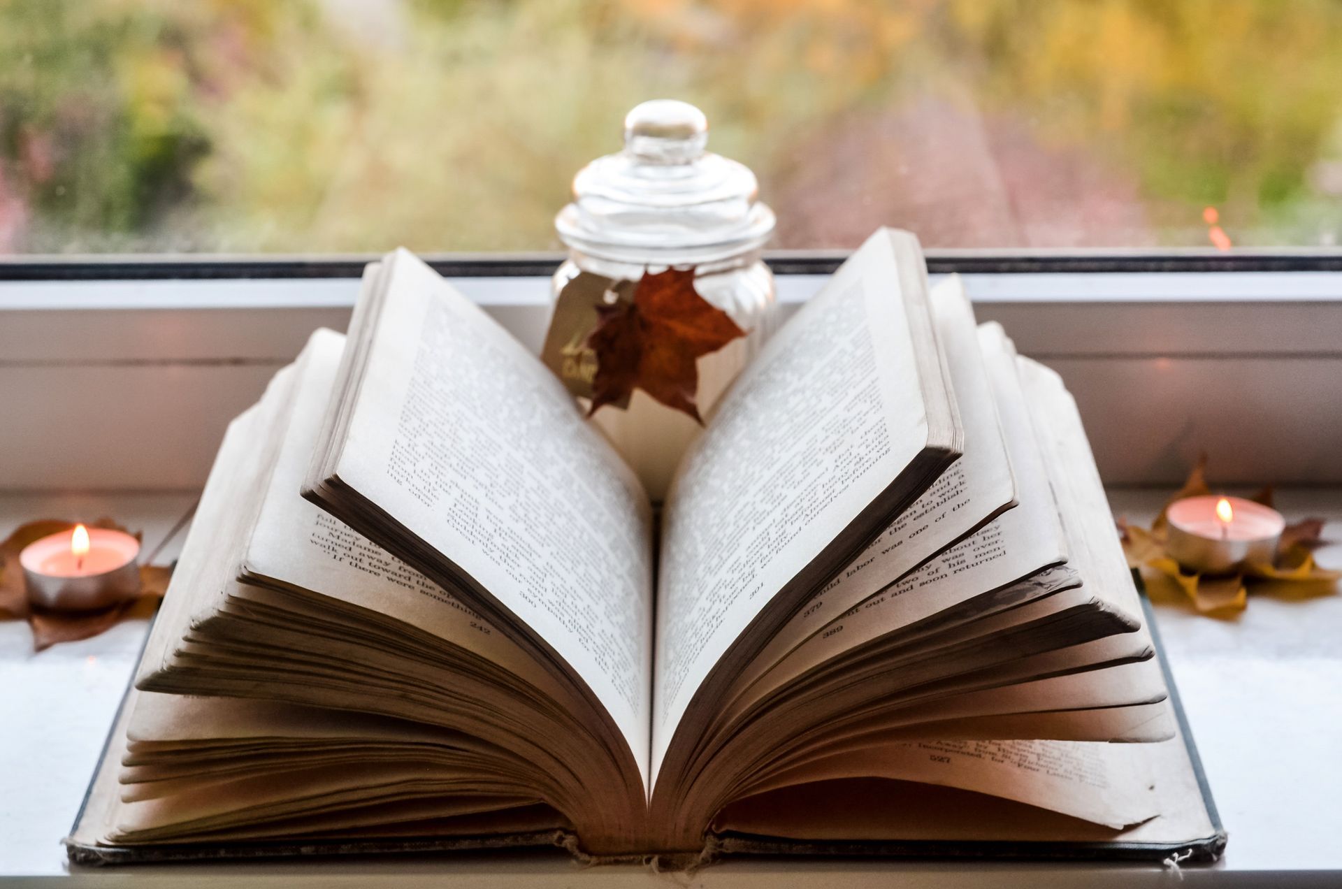 Open book on windowsill, glass jar with leaf, autumn leaves, lit candles, blurred outdoor backdrop.