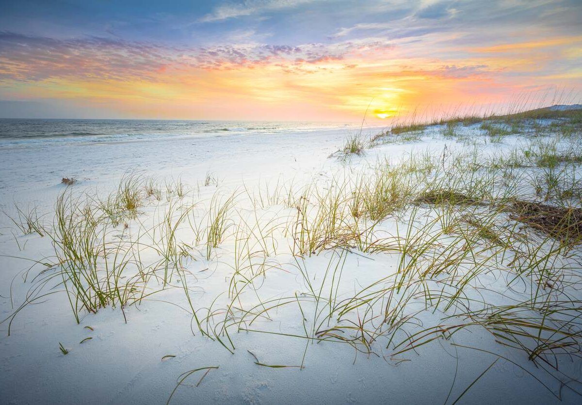 White sand beach with grassy dunes at sunset; orange and yellow sky over ocean.