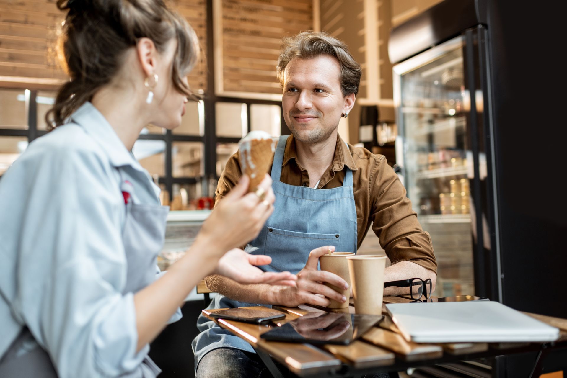 Woman and barista converse at a cafe table, holding a pastry and cups, warm setting.