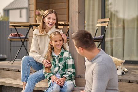 Family of three on porch, smiling. Daughter sitting between parents. Dog lying nearby.