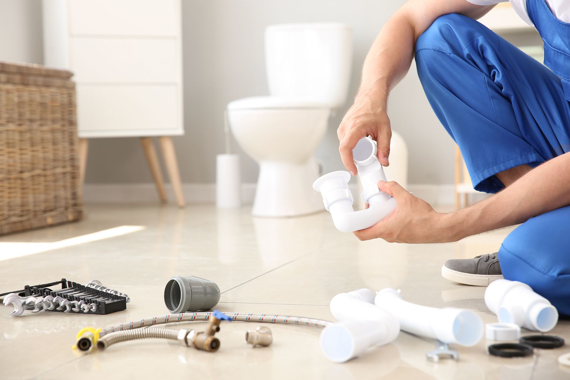 Plumber in blue overalls assembling a white drain pipe in a bathroom with a toilet and other plumbing parts.