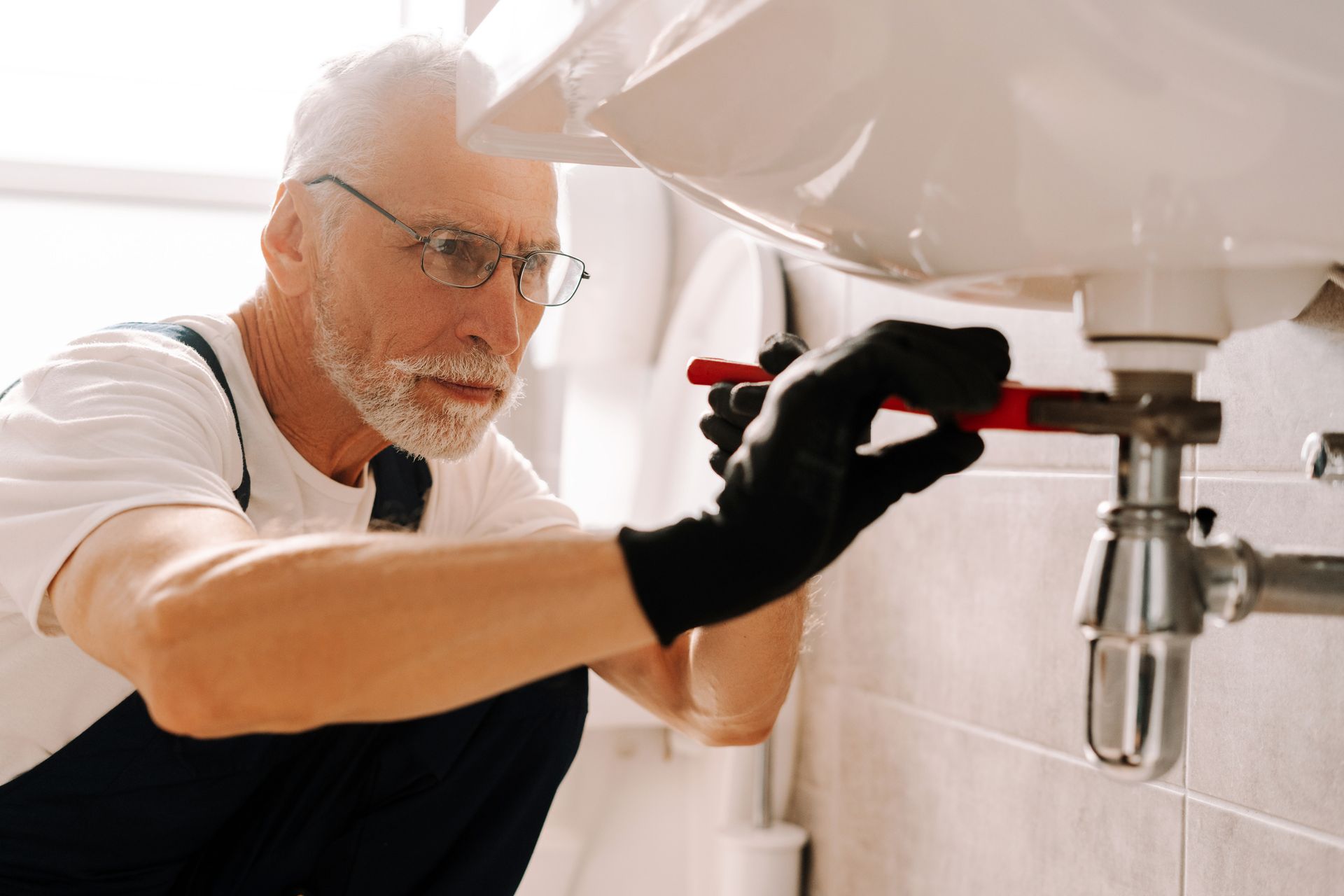 The plumber wearing gloves carefully fixes the sink pipe with adjustable pliers. The plumber wearing gloves carefully fixes the sink pipe with adjustable pliers.